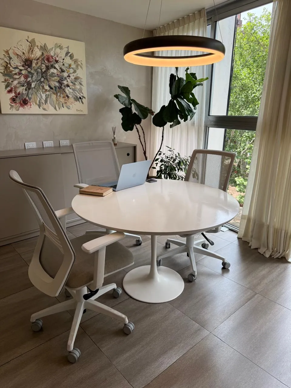 Sala de reuniones con mesa redonda blanca, cuadro floral y ventanal con plantas
